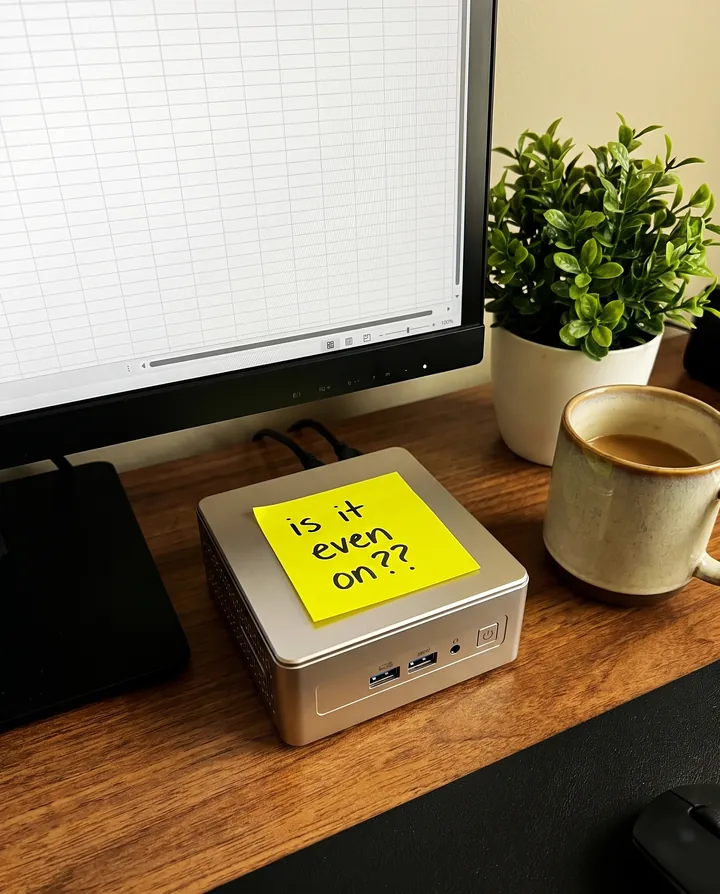 Spec ad for GEEKOM styled as a phone photo. A silver GEEKOM mini PC sits on a wooden desk next to a monitor, a plant and a coffee mug, with a yellow sticky note on top that says is it even on.