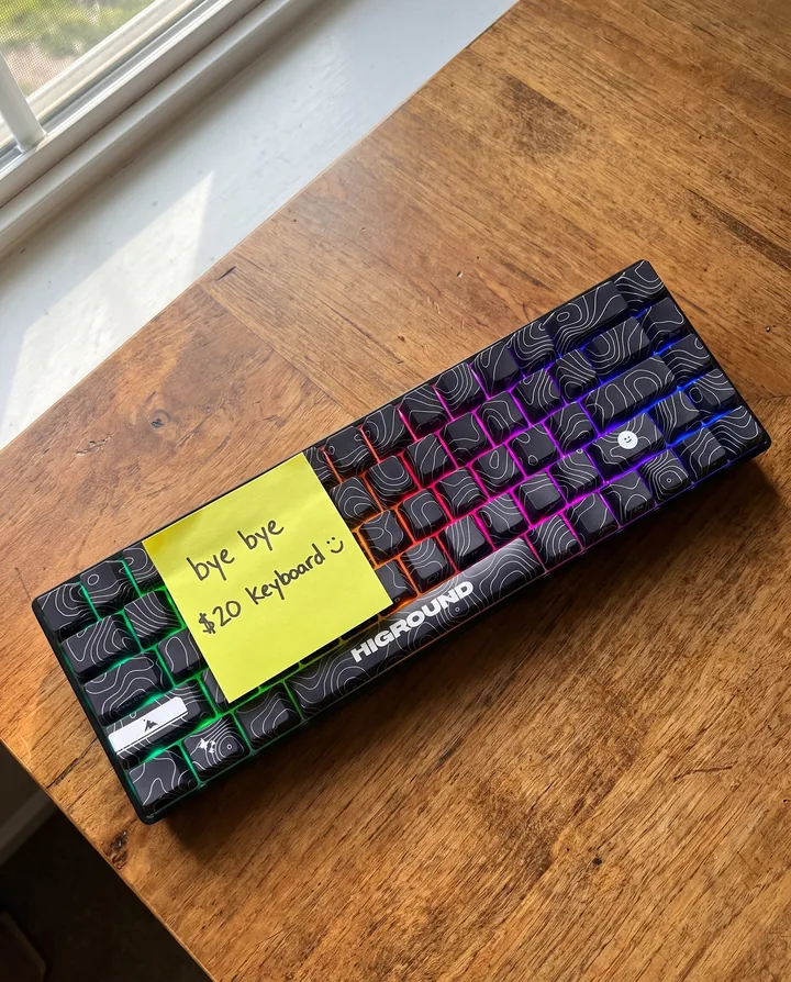 Spec ad for Higround styled as a phone photo. A glowing black Basecamp 65 BLACKICE keyboard on a wooden desk with a yellow sticky note that says bye bye $20 keyboard with a smiley.