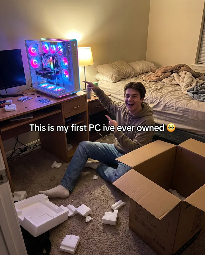Spec ad for Skytech Gaming styled as a phone photo. A grinning young man sits on his bedroom floor beside an open box and packing foam, pointing at a glowing white Skytech Prism 5 on his desk, with the caption This is my first PC ive ever owned.